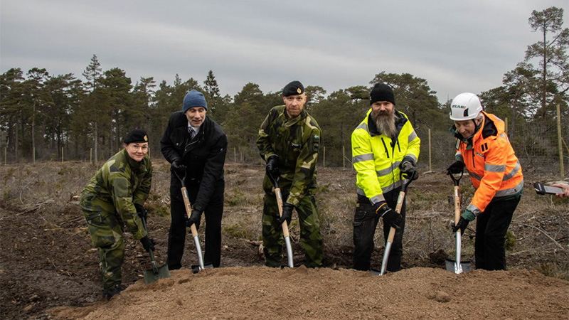 Fem personer står vända mot kameran med varsin spade i marken, redo att ta ett första spadtag.