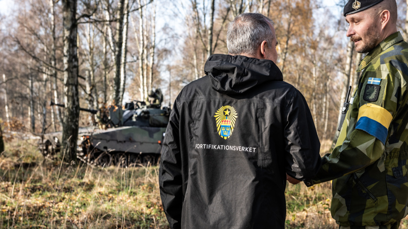 En militär och en civil med Fortifikationsverkets logotyp står i terrängen med en pansarvagn i bakgrunden