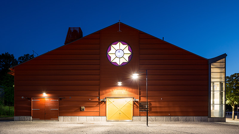 Kvällsbild av gaveln på ett stort ridhus med gul port. Över porten, i mitten av huset lyser ett runt fönster i form av en rosett.