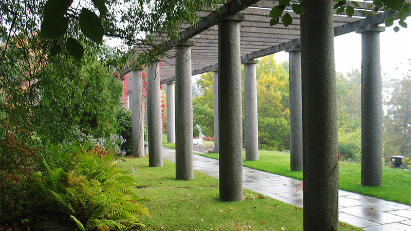 Parklandskap med en stenbelagd pergolaliknande gång med kolonner längs sidorna och takribbor av sten.