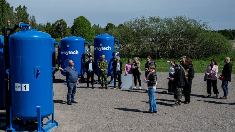 Ett antal blå cisterner står på rad och en folksamling står framför och lyssnar till en person som berättar något.