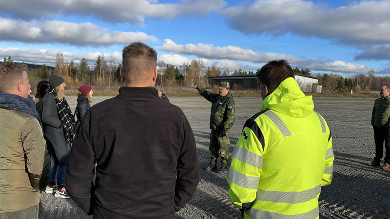 På en grusplan står en ett gäng personer och tittar mot en man som pekar och berättar. I bakgrunden syns skog och en låg byggnad.