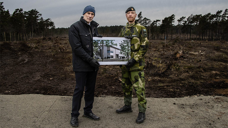 Utomhus står två män som håller upp och visar en illustrationsskiss över en byggnad.