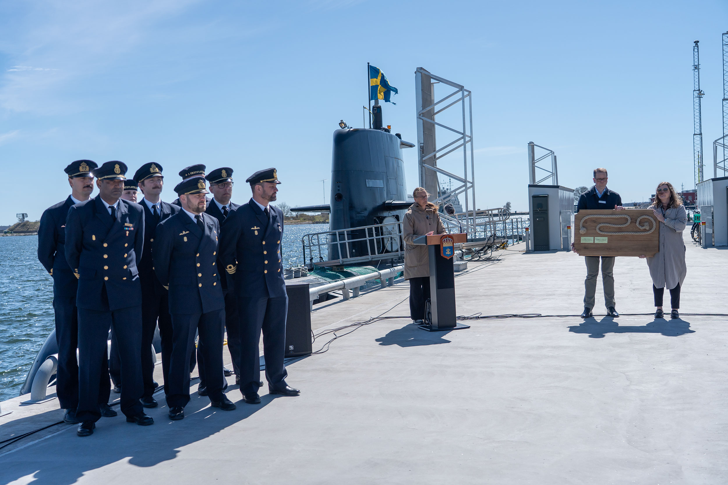 En kvinna står vid ett podium på en pir som skjuter ut i vattnet. I bakgrunden ligger en ubåt. 