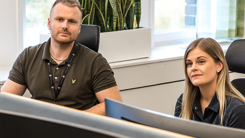 Kille och tjej lsitter vid varsitt skrivbord och yssnar när en kollega pratar.