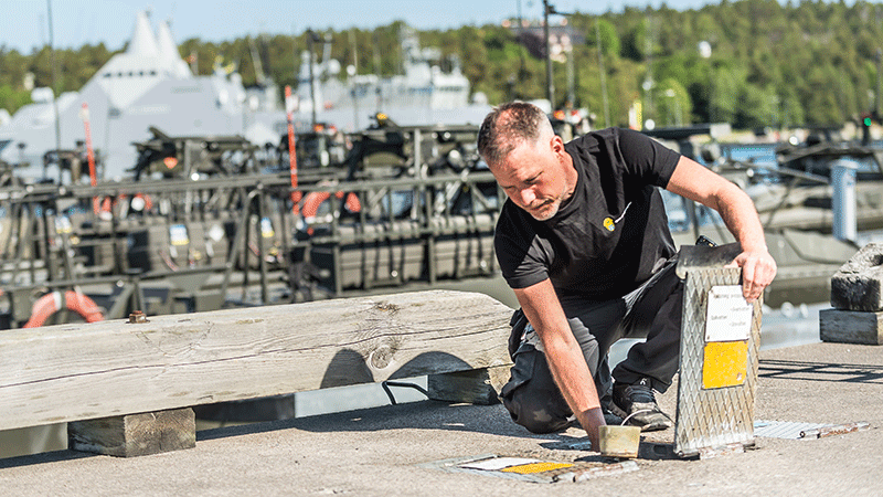 Drifttekniker sitter på knä på en betongpir och öppnar en lucka. Stridsbåtar syns i bakgrunden. 