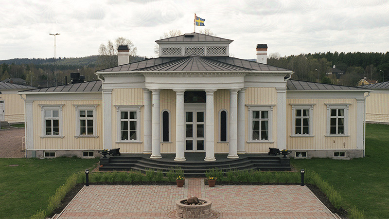 En ljusgul byggnad i nyklassicism, med vita detaljer, svart tak och en svensk flagga i topp.