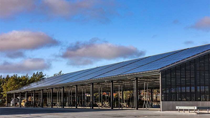 Stort skärmtak i mörk plåt med solceller på taket. Under taket står militärfordon parkerade.