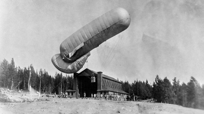 Svartvitt fotografi med en stor cigarrformad luftballong som stiger eller landar. Under syns en stor träbyggnad med portarna öppna.