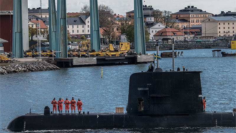 Ubåten HMS Gotland i Karlskrona hamn. På ubåten är en svensk flagga hissad och människor i orangea overaller står på däck.