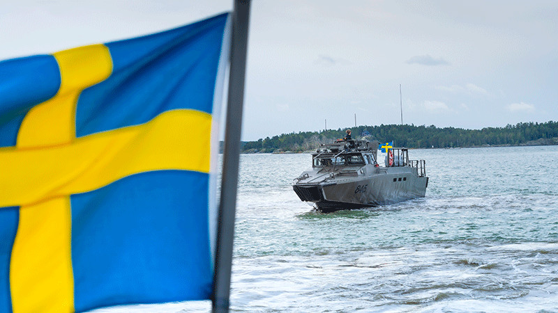 En svensk flagga vajar i förgrunden och i bakgrunden syns en stridsbåt göra sig redo att lägga till.
