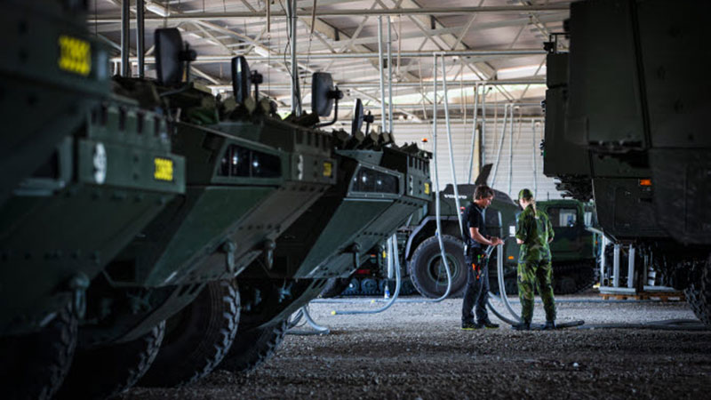 En person och en militär diskuterar en slangkoppling under ett skärmtak med stridsfordon.