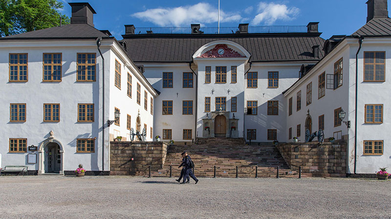 Vit slottsfasad med en trappa i mitten. Två kadetter går på en grusplan framför slottet.