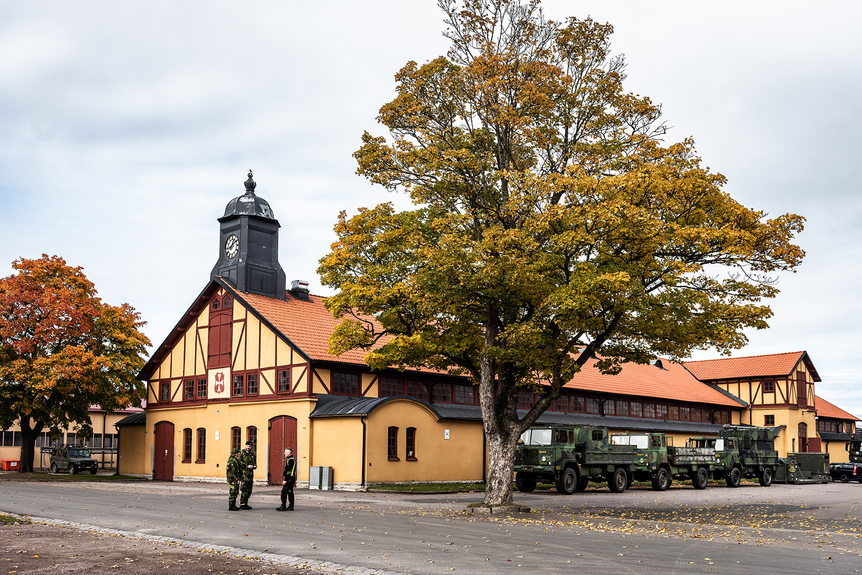 En gul och röd lång byggnad med höga träd som har löv i gula och gröna nyanser. Utanför byggnaden står tre personer och pratar med varandra.